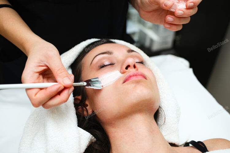 Beautician is applying a white facial mask with a brush to a